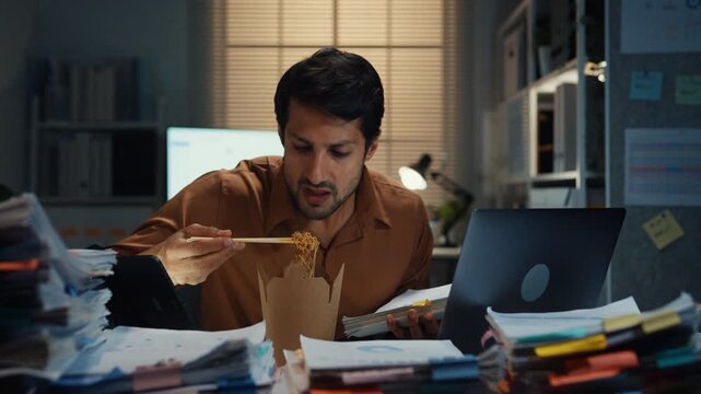 Young Latin businessman taking urgent phone call while reading accounting document on laptop at desk in dark night office. Multitasking overtime, overworked deadline, tired paperwork rush concept.
