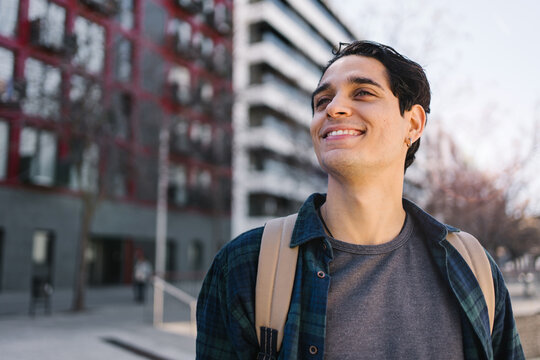 Portrait young hispanic man with backpack standing in the city looking to the side with confident expression