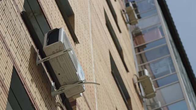 Upward view of a brick apartment building facade with an exterior air conditioner unit mounted on brackets beside an enclosed balcony, cables and piping visible in bright daylight