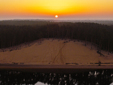 Aerial view of Lielupe White Dune at sunrise with pine trees atop the sandy slope and calm water reflecting the golden light in Jurmala, Latvia.