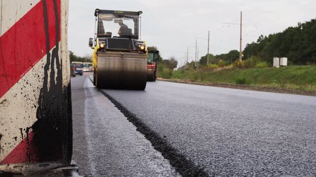 Road roller compacting fresh asphalt during highway construction and repair work with a traffic cone in the foreground