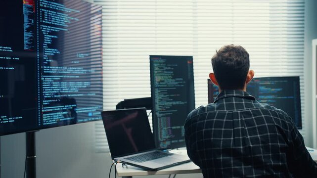 Young Caucasian male programmer writes code using multiple screens in home office workspace. AI development, cybersecurity, network database system, software engineering, remote tech startup concept.