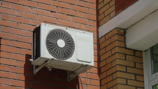 Outdoor air conditioning condenser unit mounted on a brick building facade near a window, showing circular fan grille, metal support brackets, and housing under bright daylight