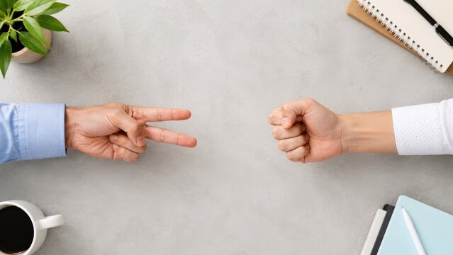 Two hands playing rock paper scissors on a desk with coffee, notebook and a small potted plant