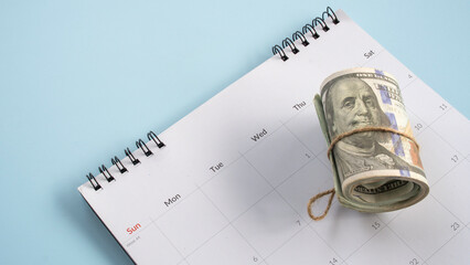 Roll of US dollar bills sitting on top of a monthly desk calendar planner concept of financial deadlines tax day payment schedule or savings © MohamadFaizal