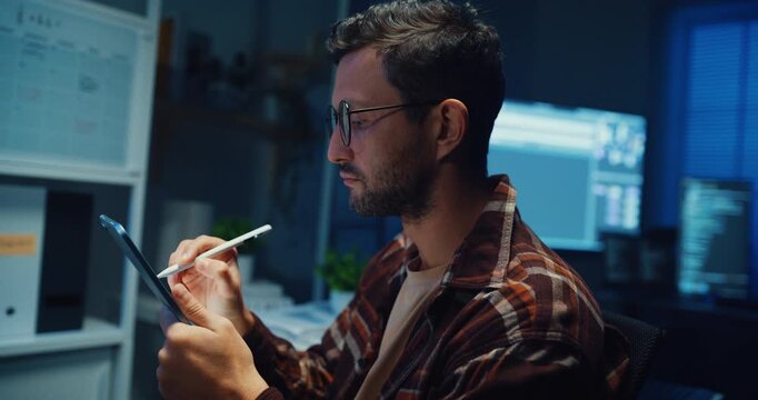 Young Caucasian male programmer use tablet for code design and plan in dark home office at night. AI technology, cybersecurity, software developer, freelance innovation, digital data network concept.