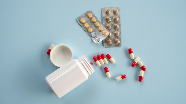 A flat lay of a white pill container with scattered red and white capsules alongside silver blister pack medication strips on a light background.