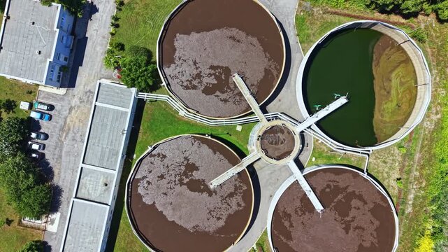 Workers manage the treatment of wastewater in three large round tanks, while vehicles are parked nearby and green areas surround the facility.
