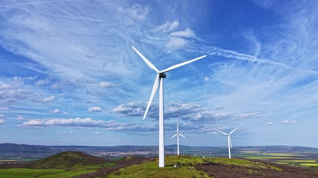 Three wind turbines rotate gently on a hillside covered in grass and small shrubs. The sky is bright with clouds, and fields extend into the distance.