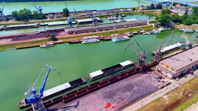 Drone view of industrial harbor cranes, Bratislava, Slovakia. High angle shot of blue loading machinery and cargo barges along the green riverbank.
