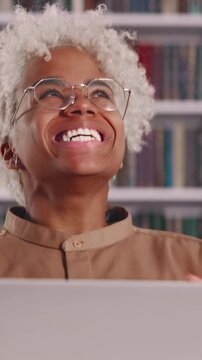 Excited young African American student with glasses gasps, cheers, and celebrates success in front of her laptop, smiling widely and expressing pure happiness inside a library.