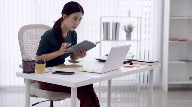 Young Asian businesswoman referencing notebook while typing on laptop during focused work session at modern home office