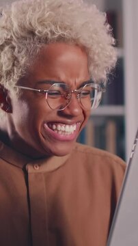 Excited young African American student wearing glasses gasps, cheers, and celebrates a great success on her white tablet, smiling widely and expressing deep joy in her library.