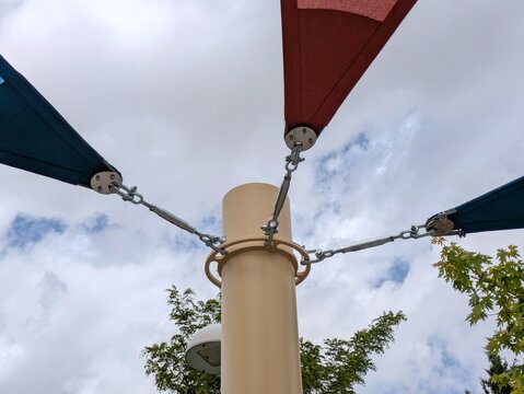 Vista de cerca de tensores con cables de metal en la parte superior de postes de sujeci&oacute;n de parasoles triangulares de exterior, de color rojo y azul, en plaza p&uacute;blica, con cielo con nubes de fondo 