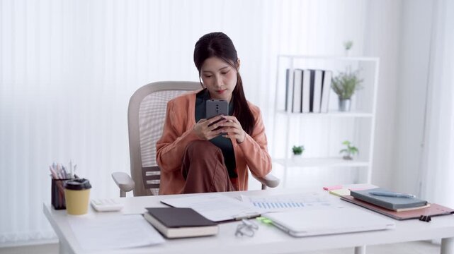 Young Asian professional woman reviewing business reports on smartphone while analyzing financial data at modern home office desk