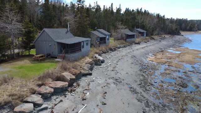 Drone footage gliding along rustic waterfront cabins beside a rocky tidal shoreline, bordered by dense forest and calm coastal waters.