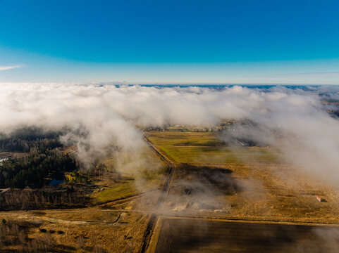 Aerial view of fields, forest, and the A8 highway partially covered by low-hanging clouds under a clear blue sky in Olaine parish, Olaine Municipality, Latvia.
