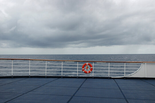 An empty cruise ship deck with a lifebuoy ring on the railing faces an open sea under heavy grey storm clouds