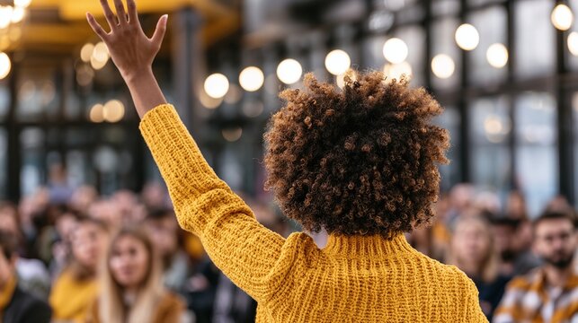 Rear view of a confident individual with curly hair and a yellow sweater, hand raised, addressing an audience in a bright, modern setting, conveying engagement and interaction