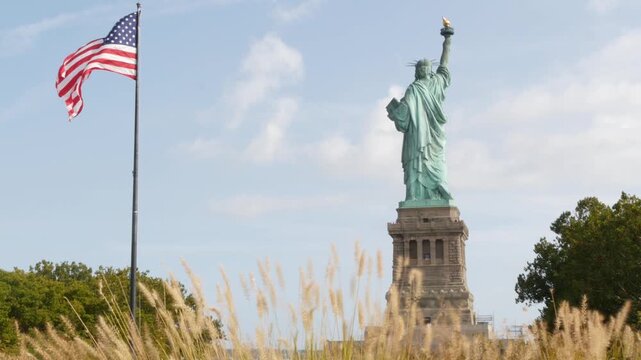 Statue of Liberty on Liberty Island, New York, United States. Cooper monument from behind, democracy and patriotism symbol near Manhattan, NYC USA. American flag waving in wind. Independence concept.