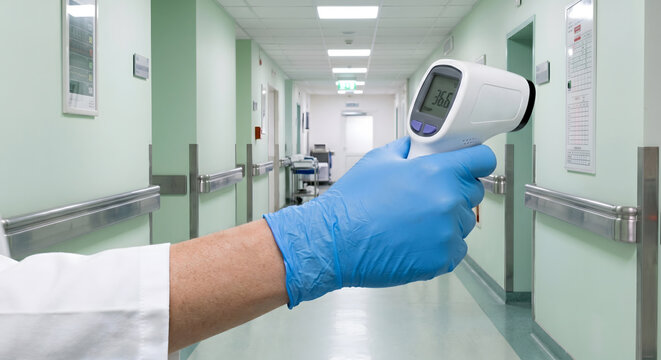 Medical professional hand wearing blue glove holding digital infrared thermometer in hall.