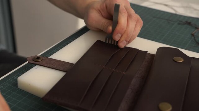 Leather craftsman using stitching chisel and mallet on brown wallet in his workshop, creating handmade accessory