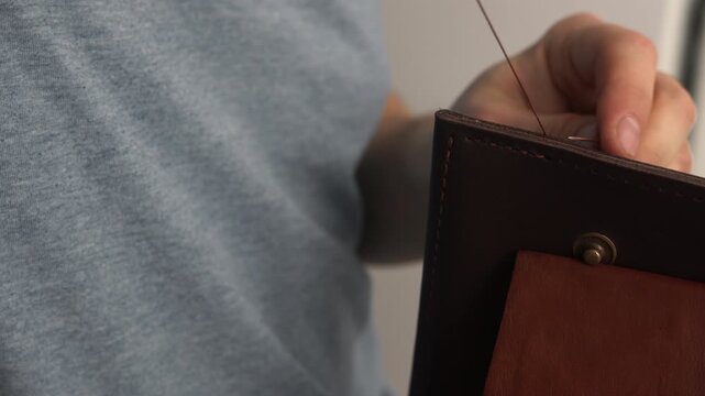 Leather craftsman hand sewing a brown leather wallet using traditional saddle stitching technique in a workshop