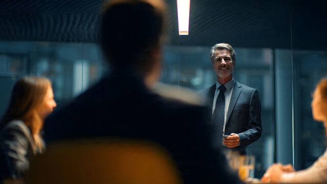 A mature confident male businessman stands leading a corporate boardroom meeting and presentation for colleagues focusing on effective teamwork collaboration inside a modern workplace environment.