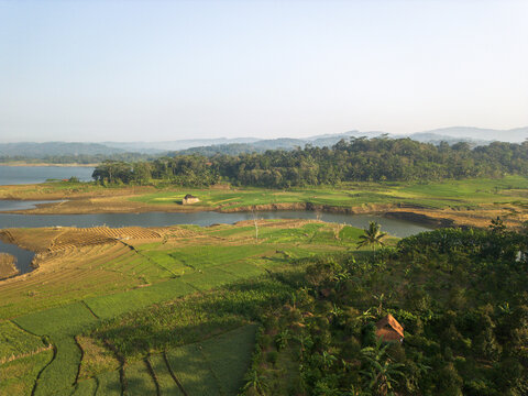 Aerial view of Wadaslintang Reservoir surrounded by lush green agricultural fields, terraced crops, and dense tropical forests in Wadaslintang, Central Java, Indonesia.