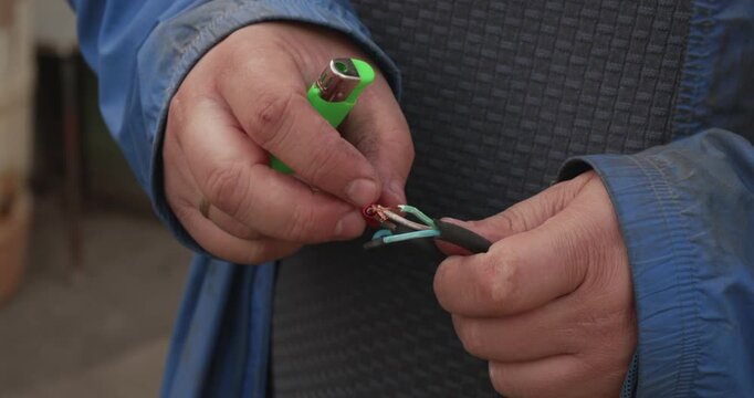 Electrician's hands are repairing a damaged electrical cable by twisting the copper wires and using a lighter to apply heat shrink tubing for insulation on each conductor.