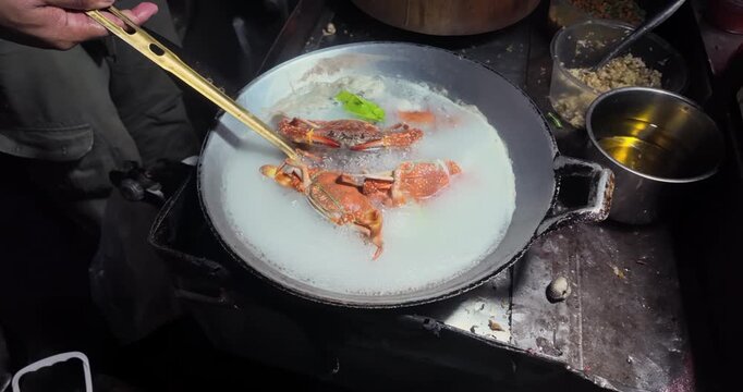 Close up: at night, a cook uses a strainer to stir crabs boiling in a milky broth inside a wok.