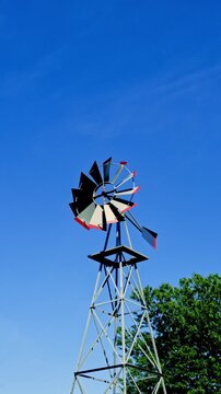A windmill spins slowly in the breeze under a clear blue sky. Trees are nearby, offering shade on a bright sunny day. The scene shows a typical rural setting with few clouds in the sky.
