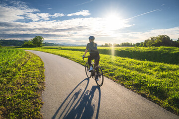 Fototapeta premium Female cyclist with road bike riding on asphalt path at sunset in summer. Slovenia. Rear Woman cycling in helmet on bicycle lane near green fields, active sport lifestyle, sun sky landscape. Lifestyle