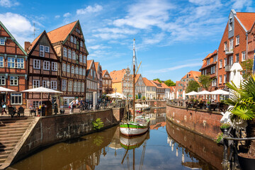 Obraz premium Historic harbor of Stade with half timbered houses, restaurants, visitors and traditional sailing vessel beneath blue sky with reflections in the water.