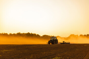 Fototapeta premium Tractor Working in Field with Dust Cloud at Sunset Golden Hour