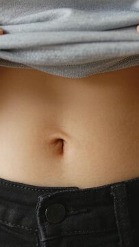 Vertical video: Hands lifting grey top hem, revealing belly and pinching navel over jeans at home