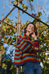 Fototapeta premium Woman in a colorful striped sweater leaning on a bamboo trellis outdoors, smiling and thoughtful in a casual outfit with jeans in an autumn garden lifestyle and fashion portrait.