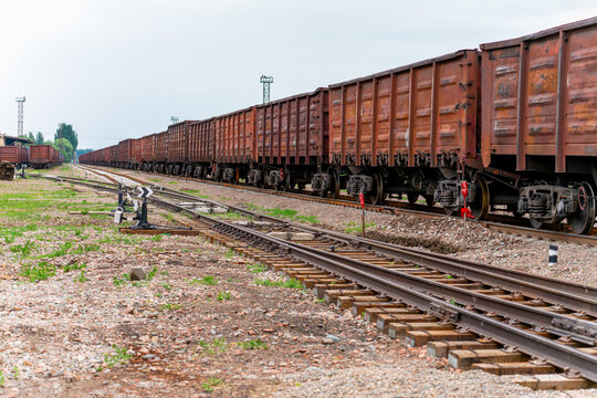 A long industrial freight train consisting of rusty brown open high-sided wagons stands on the tracks of a sorting yard under a cloudy sky. 