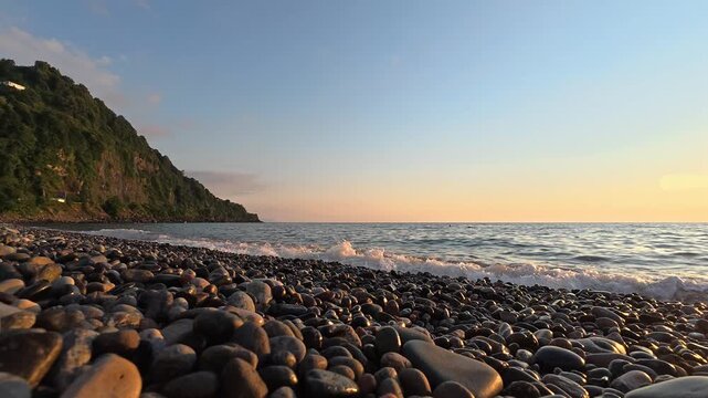 4K footage of gentle sea waves washing over a pebble beach during sunset. Scenic coastline with rocky cliffs and golden evening light in Adjara, Georgia.