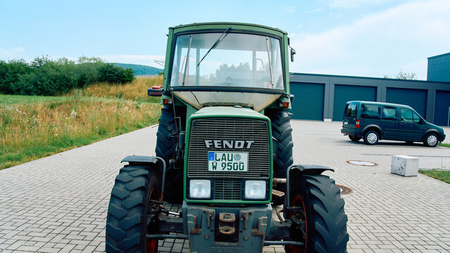 Front view of a classic green Fendt tractor with a German license plate, parked on a paved lot with a van and hills behind.