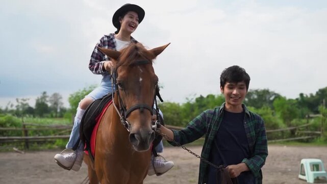 Near-end sequence frame shows smiling woman riding brown horse while young man guides bridle beside her in rural paddock with fence, trees, and soft daylight