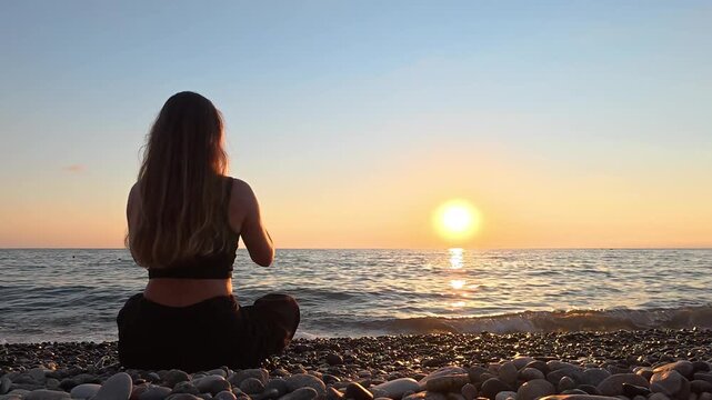 4K footage of a young woman sitting on a pebble beach and watching the sunset over the Black Sea. Peaceful evening atmosphere, mindfulness, meditation and relaxation concept in Georgia.