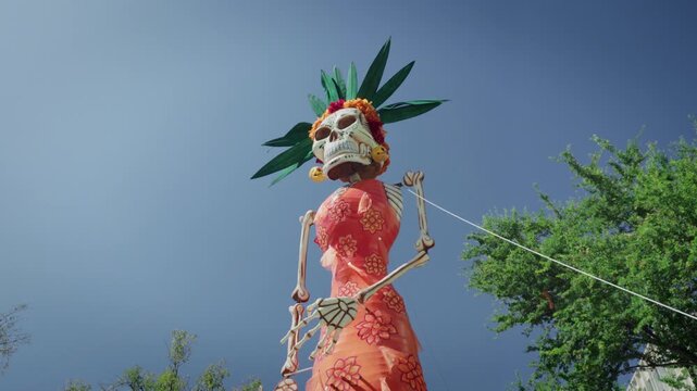 Handheld shot captures colorful Day of the Dead street decorations in Zapopan, Jalisco, with skeleton figures and elegant Catrina sculptures adorning the avenue against lush green trees and a vivid bl