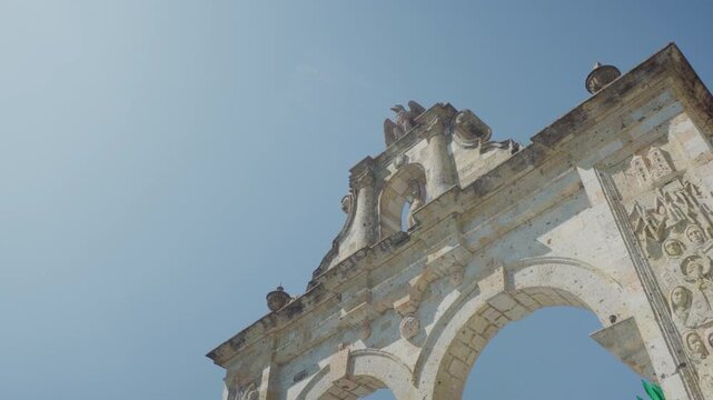Aerial drone shot looking up at the Zapopan Arch, capturing its decorative columns and eagle sculpture at the top against a vivid blue sky in Guadalajara, Jalisco