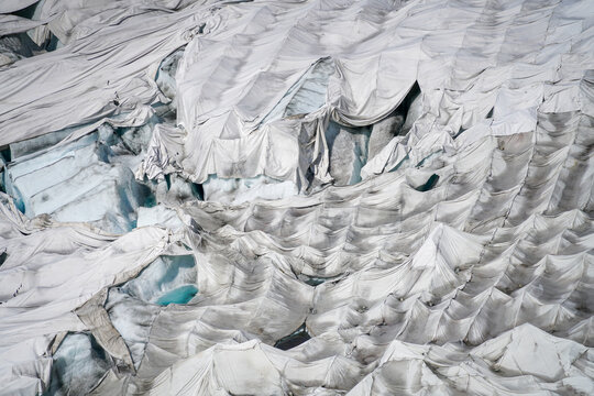 Aerial view of the Rhone Glacier covered with white protective tarps to prevent melting, showing ice crevices and blue meltwater pools in Obergoms, Valais, Switzerland.