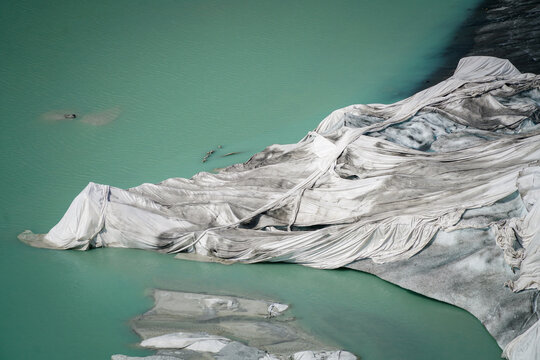 Aerial view of the Rhone Glacier covered with protective white fleece to reduce melting, situated beside a turquoise glacial lake in Obergoms, Valais, Switzerland.