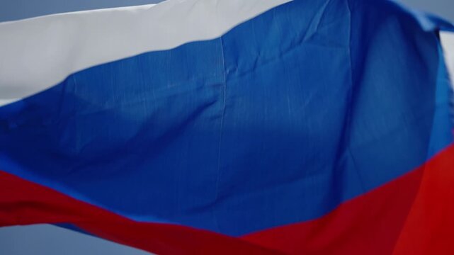 Close-up of a white, blue, and red horizontal tricolor flag billowing in the wind against a clear blue sky, captured across two video frames with folds, motion, and daylight.
