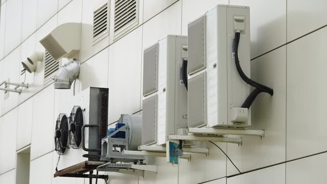 Row of external air conditioning condensers and ventilation fans mounted on a modern building facade, showing HVAC outdoor units with ducts, brackets, and weathered installations
