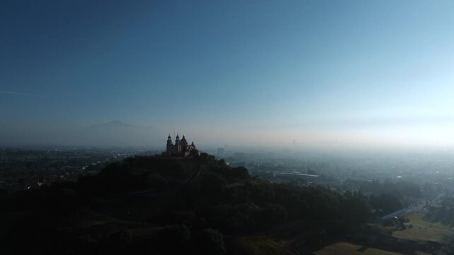 Aerial drone orbits the hilltop church atop the Cholula pyramid at dawn, with the Puebla skyline spreading below and the Popocatepetl and Iztaccihuatl volcanoes rising on the horizon