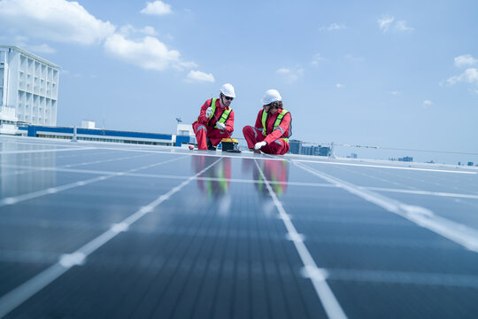 A field engineer in red coveralls and a hard hat crouches low to inspect a solar module overlooking the city, showing sustainable power, inspection and maintenance on a rooftop photovoltaic.